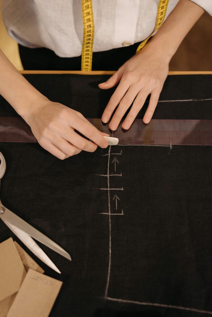 Hands marking fabric with chalk for dressmaking. Includes measuring tape and scissors.